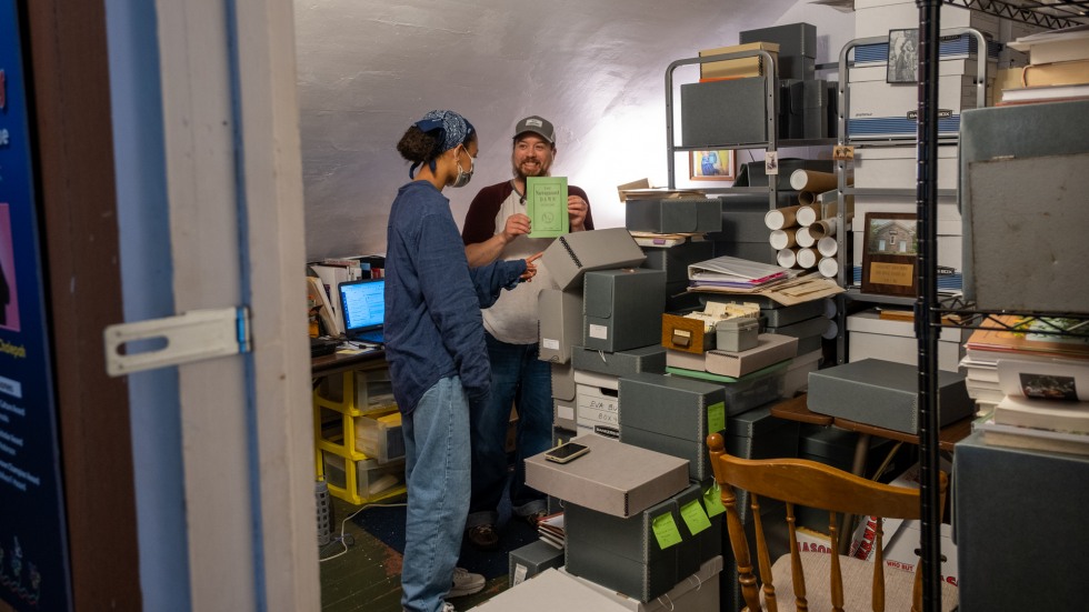 archivist holding up a copy of the Narragansett Dawn in a room full of boxes and folders